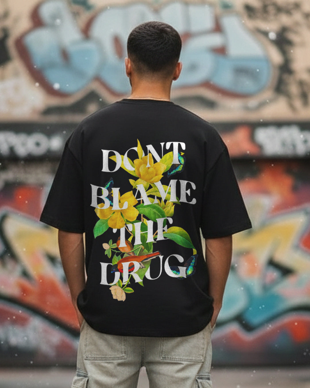 Back view of a man in an urban setting wearing a black oversized t-shirt with the white text "DON'T BLAME THE DRUG" intertwined with yellow flowers, birds, and butterflies.