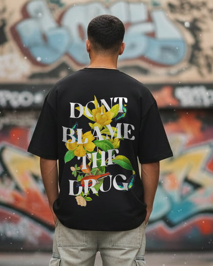 Back view of a man in an urban setting wearing a black oversized t-shirt with the white text "DON'T BLAME THE DRUG" intertwined with yellow flowers, birds, and butterflies.