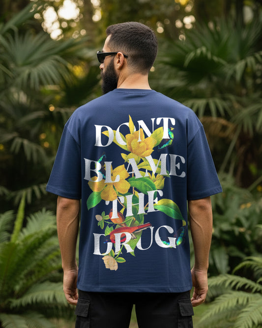 Back view of a 240 GSM navy blue oversized t-shirt featuring the white text "DON'T BLAME THE DRUG" layered with detailed illustrations of yellow flowers, tropical birds, and butterflies.
