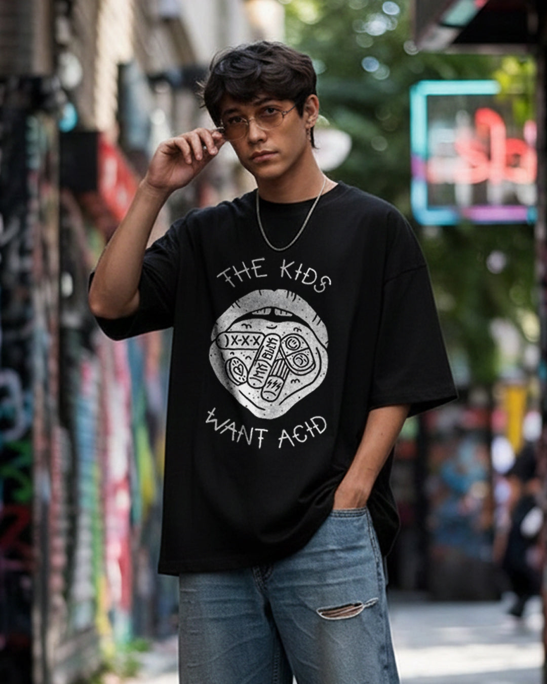 Front view of a male model in an urban alley wearing 'The Kids Want Acid' black oversized t-shirt, featuring a white graphic of a mouth with tabs on the tongue and bold typography.