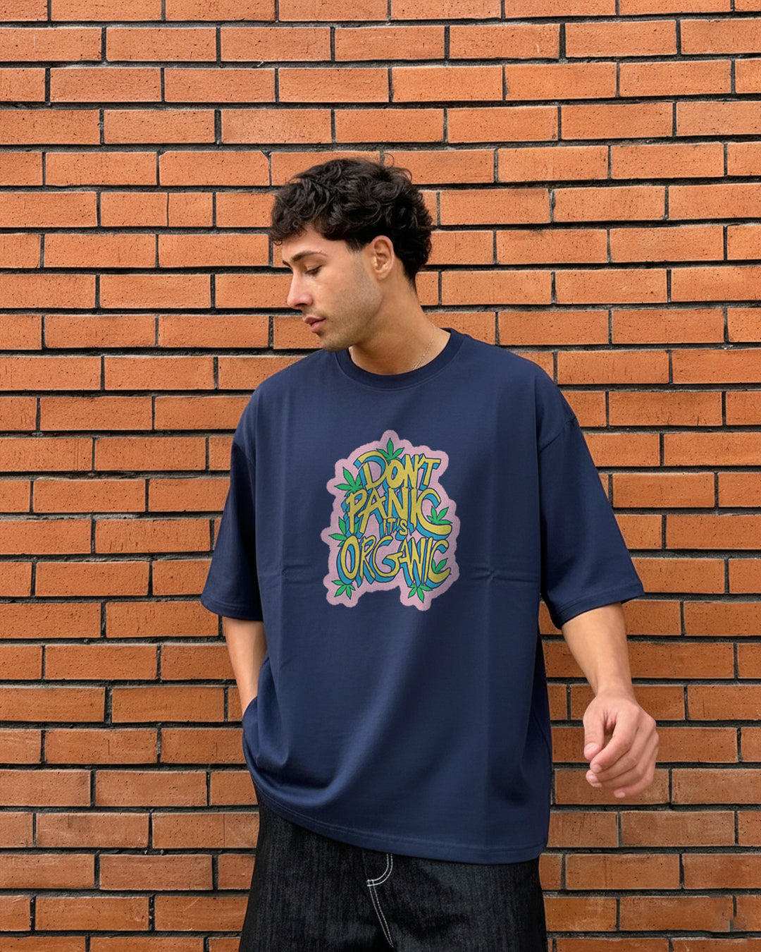 Front view of a man in front of a brick wall wearing an oversized navy blue t-shirt with a colorful graffiti-style graphic reading "DON'T PANIC IT'S ORGANIC" decorated with green leaves.