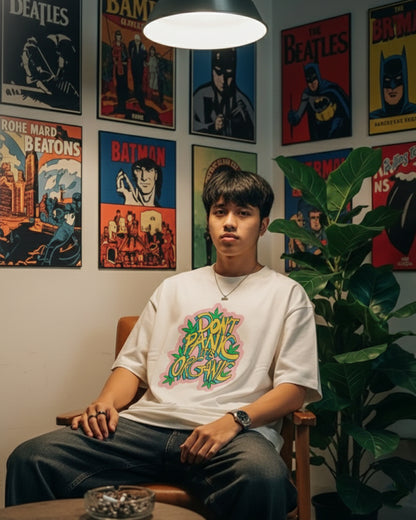 Front view of a man sitting in a relaxed position wearing an off-white oversized t-shirt with a colorful graffiti-style graphic reading "DON'T PANIC IT'S ORGANIC" decorated with green leaves.