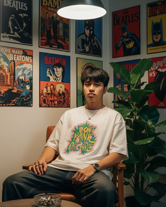Front view of a man sitting in a relaxed position wearing an off-white oversized t-shirt with a colorful graffiti-style graphic reading "DON'T PANIC IT'S ORGANIC" decorated with green leaves.