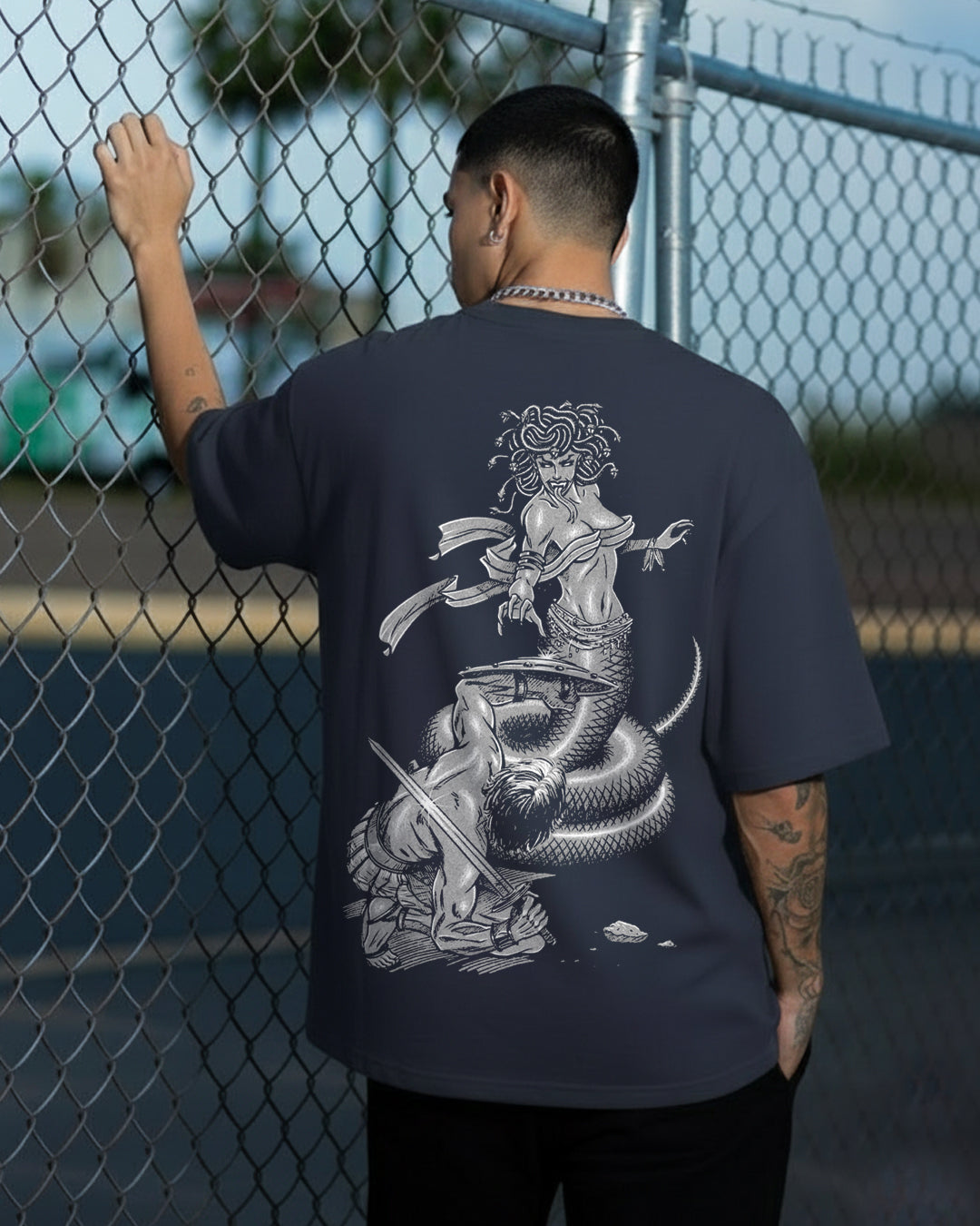 Back view of a navy blue oversized t-shirt featuring a detailed monochromatic graphic of Medusa with snakes for hair fighting a warrior with a sword and shield.