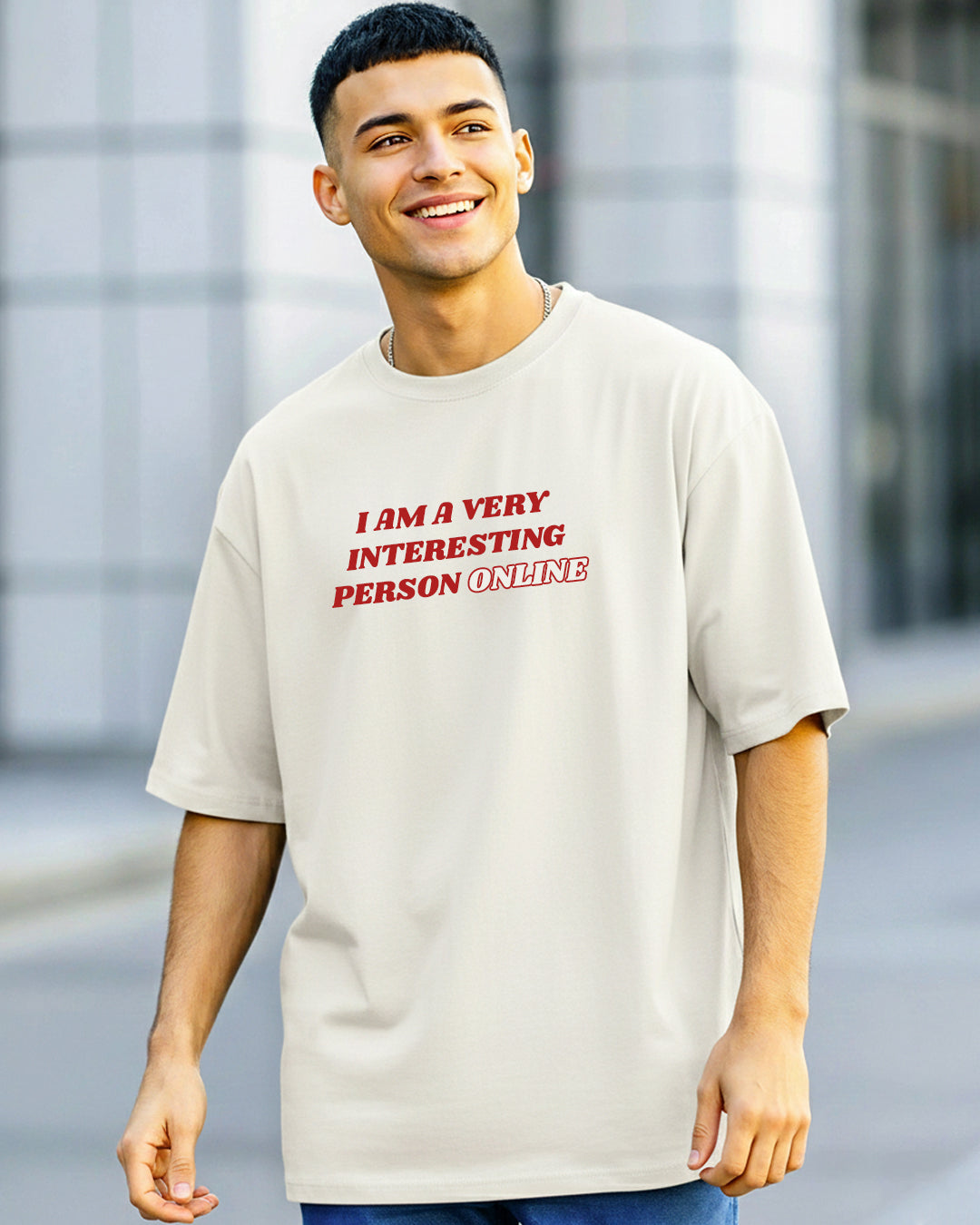 Front view of a smiling man in an urban setting wearing an off-white oversized t-shirt with the red text 'I AM A VERY INTERESTING PERSON ONLINE' printed on the chest.
