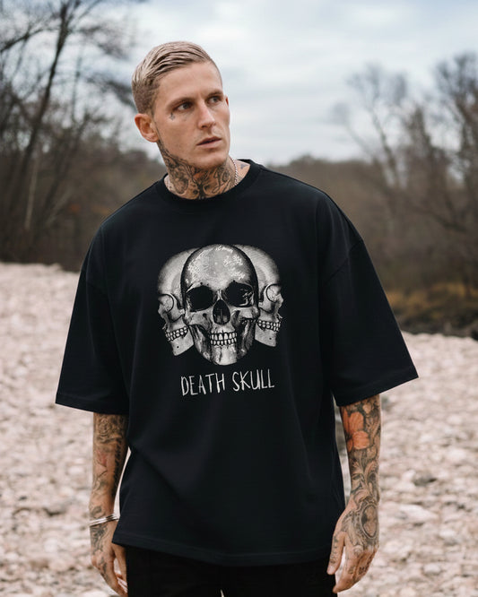 Front view of a heavily tattooed man wearing the 'Triple Death Skull' oversized black t-shirt featuring a monochromatic graphic of three skulls and the text 'DEATH SKULL'.